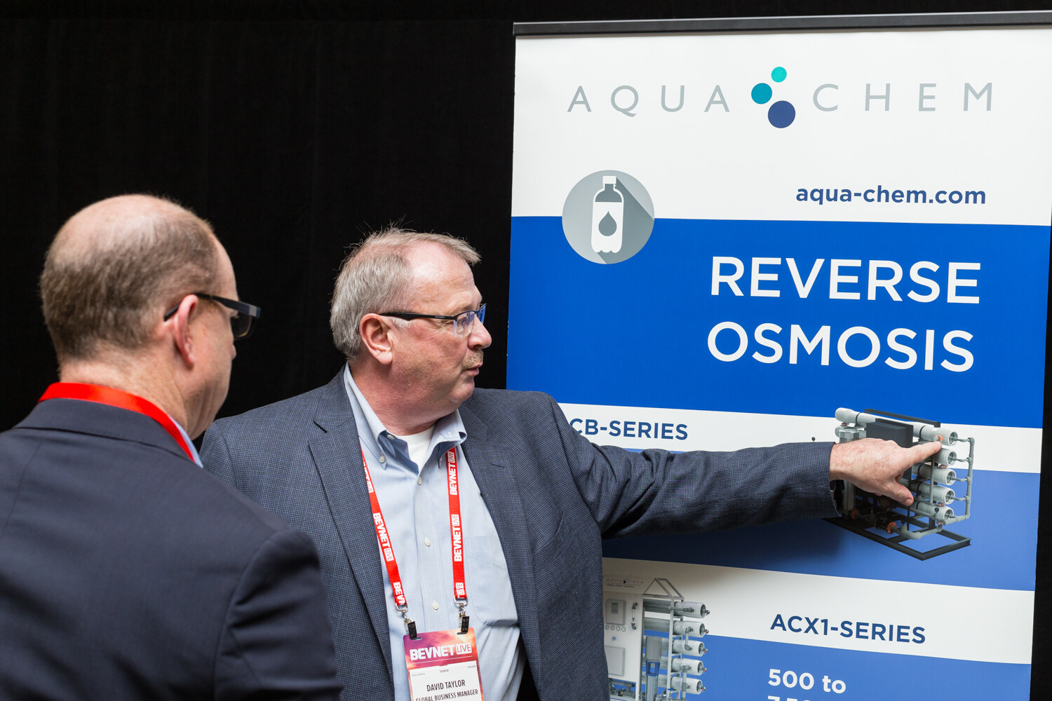 A man in a gray blazer and glasses points at a banner reading Aqua-Chem Reverse Osmosis with ACX1-Series equipment pictured, while a bald man in glasses seen from behind watches.