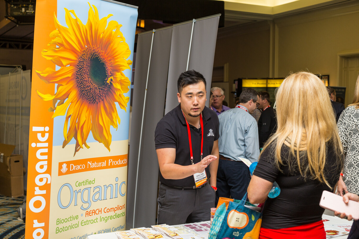 A man in a black polo and red lanyard gestures while speaking with a blond woman seen from behind, beside a sunflower banner reading Draco Natural Products, Certified Organic.