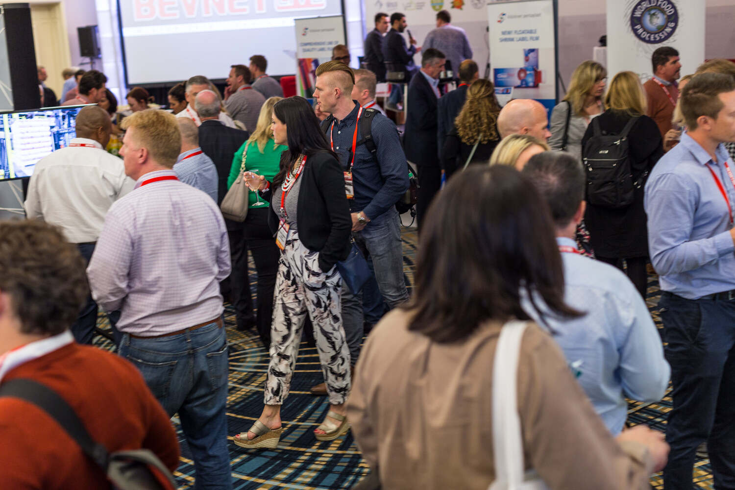 A busy crowd of attendees with red lanyards stands and walks across a patterned carpet between booths, with a World Food Processing banner and the BevNET Live stage screen in the background.