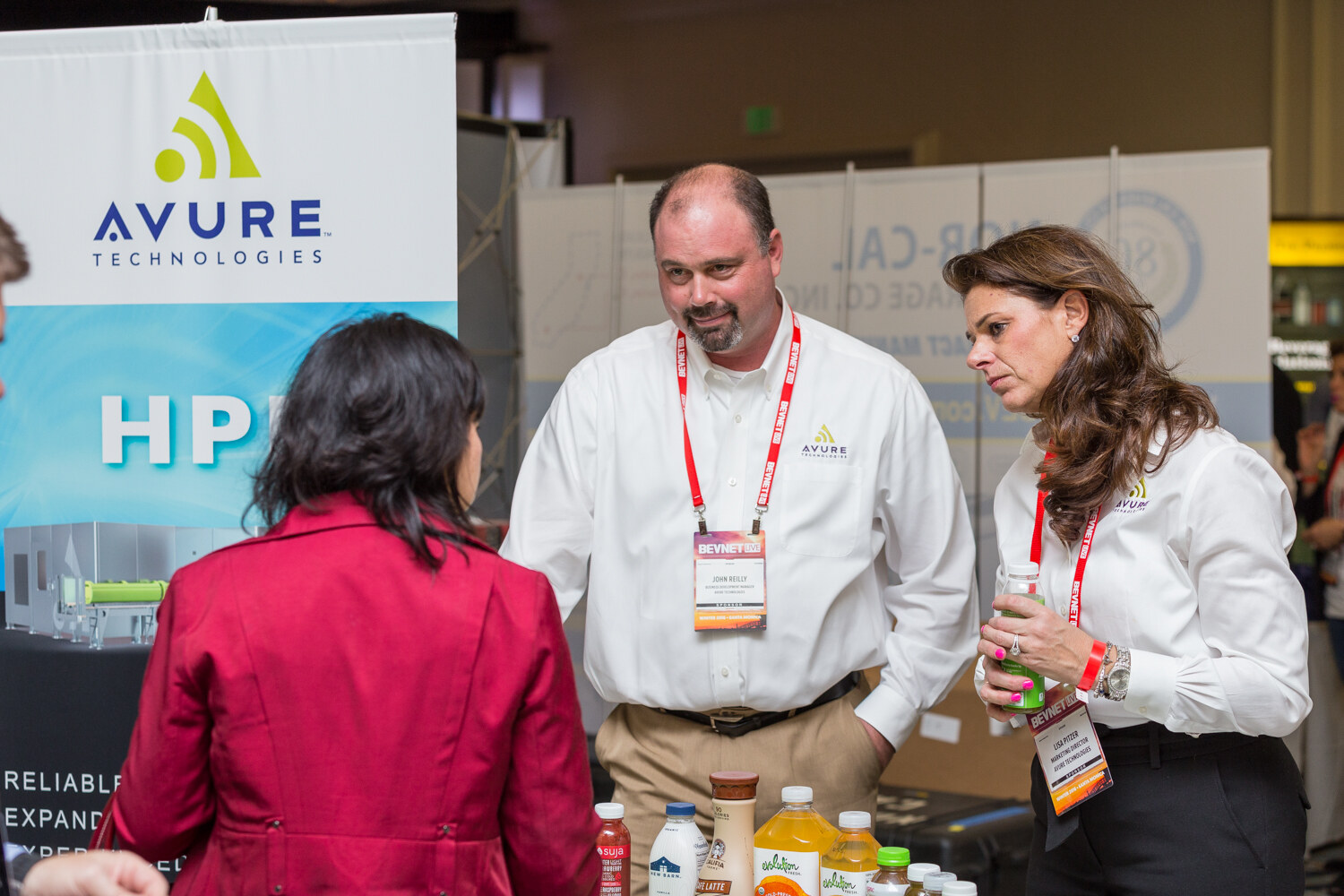A bearded man and a woman, both in white Avure shirts with BevNET Live badges, listen to a visitor in a red jacket seen from behind, beside an Avure Technologies HPP banner and sample bottles.