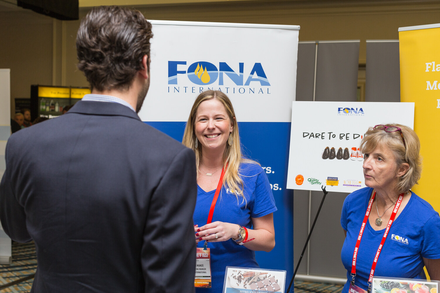 A smiling woman and an older woman in blue FONA International shirts stand in front of a FONA International banner and a Dare to be different sign while a man in a dark blazer faces them.