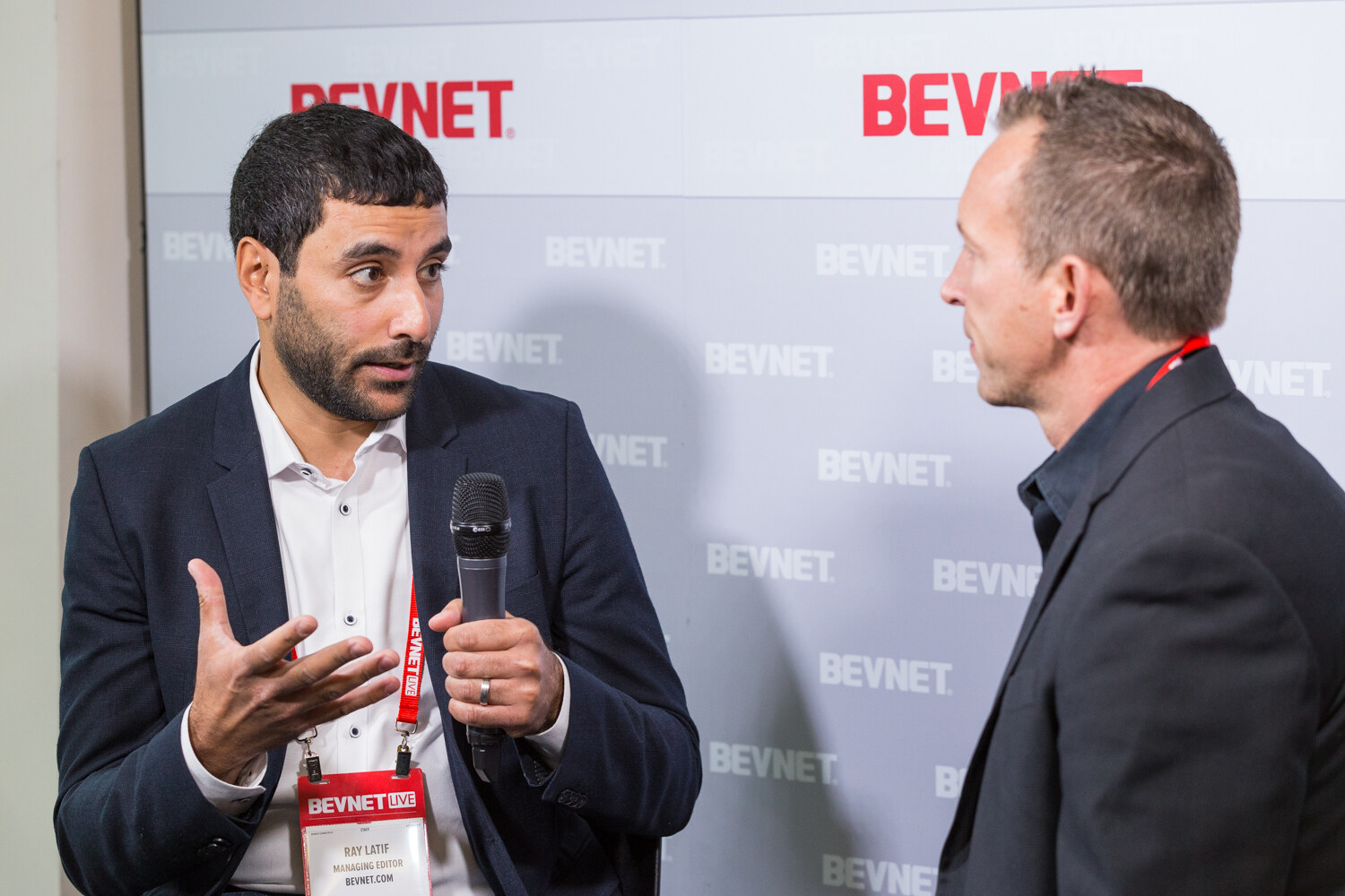 A bearded man in a navy blazer and BevNET Live badge gestures with one hand while holding a microphone and speaks with a man in a dark jacket in profile, with BEVNET logos on the wall behind them.