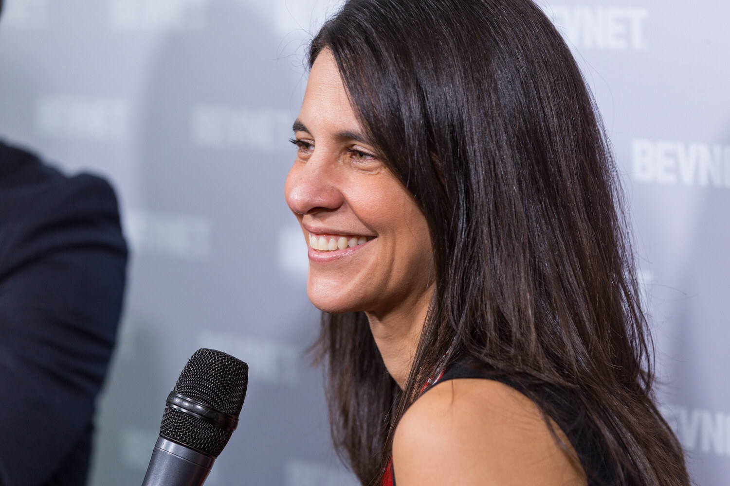 Close view of a woman with long dark hair in profile smiling, with a black microphone in the foreground and a BEVNET logo wall behind her.