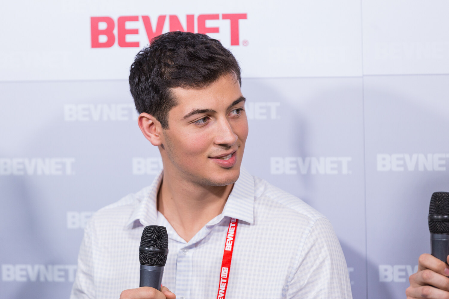 A man with short dark hair in a white checked shirt and a red BevNET Live lanyard holds a black microphone in front of the BEVNET logo wall.