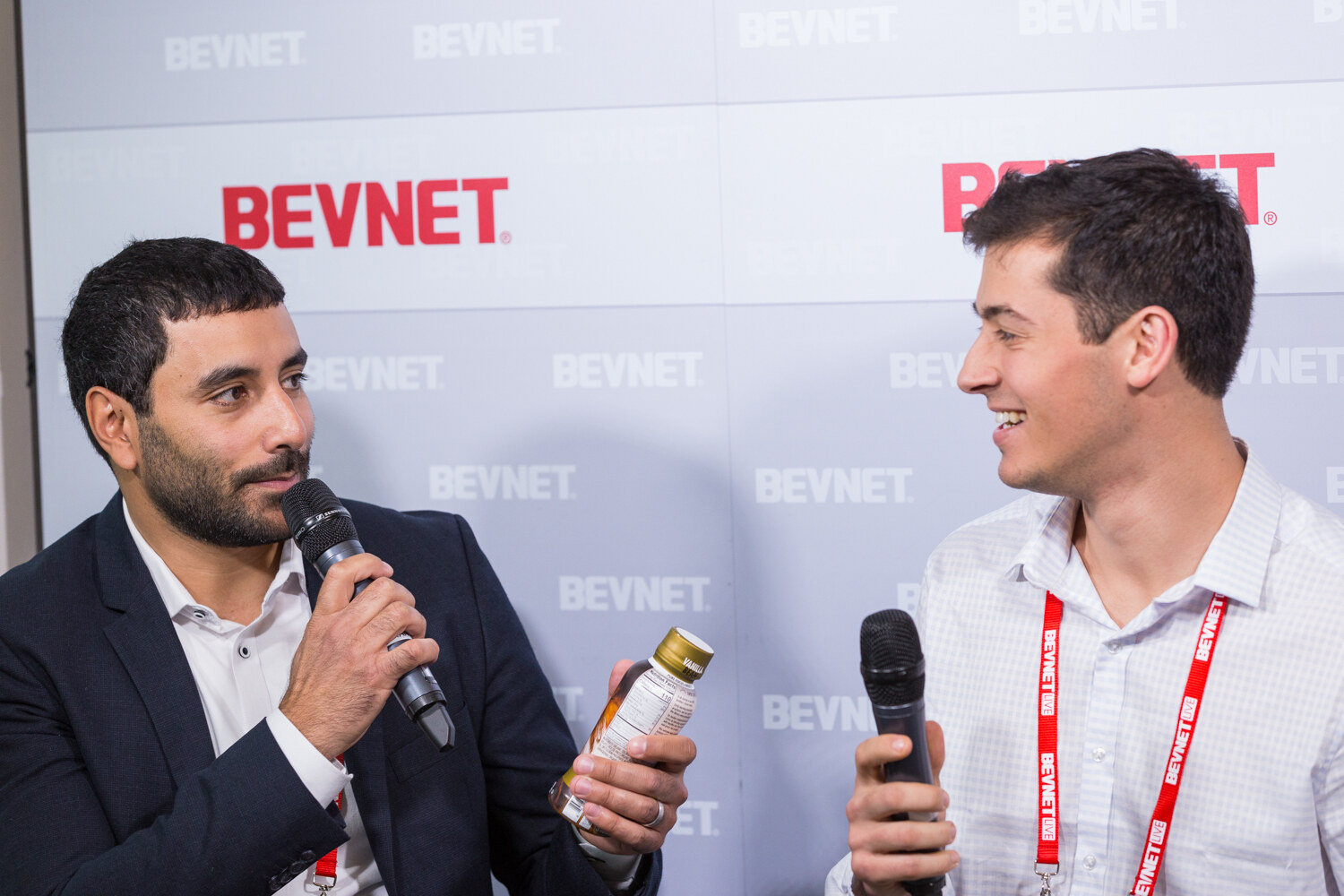 A bearded man in a navy blazer holds a microphone and a small bottle with a gold cap while a smiling man in a white checked shirt holds another microphone, in front of a BEVNET logo wall.