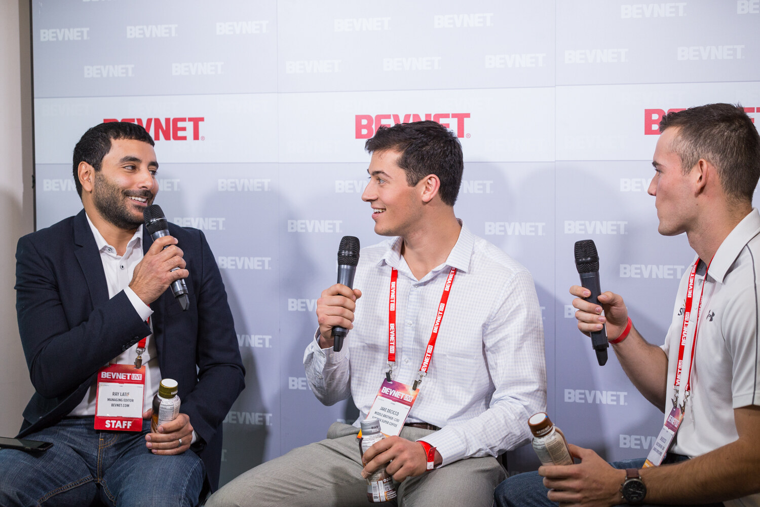 Three men sit on stools in front of a BEVNET logo wall holding microphones and small coffee bottles; the man at right wears a white Under Armour polo.