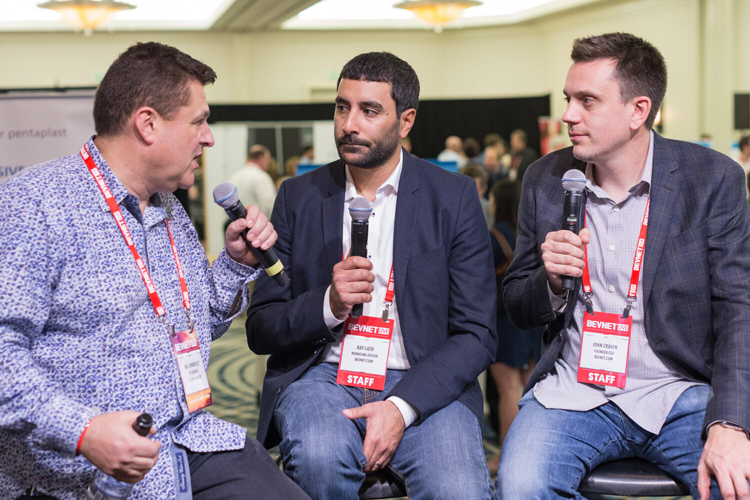 Three men wearing BevNET Live badges sit on stools holding handheld microphones: a man in a blue patterned shirt at left, a man in a navy blazer in the middle and a man in a gray plaid blazer at right, with the blurred expo hall behind them.
