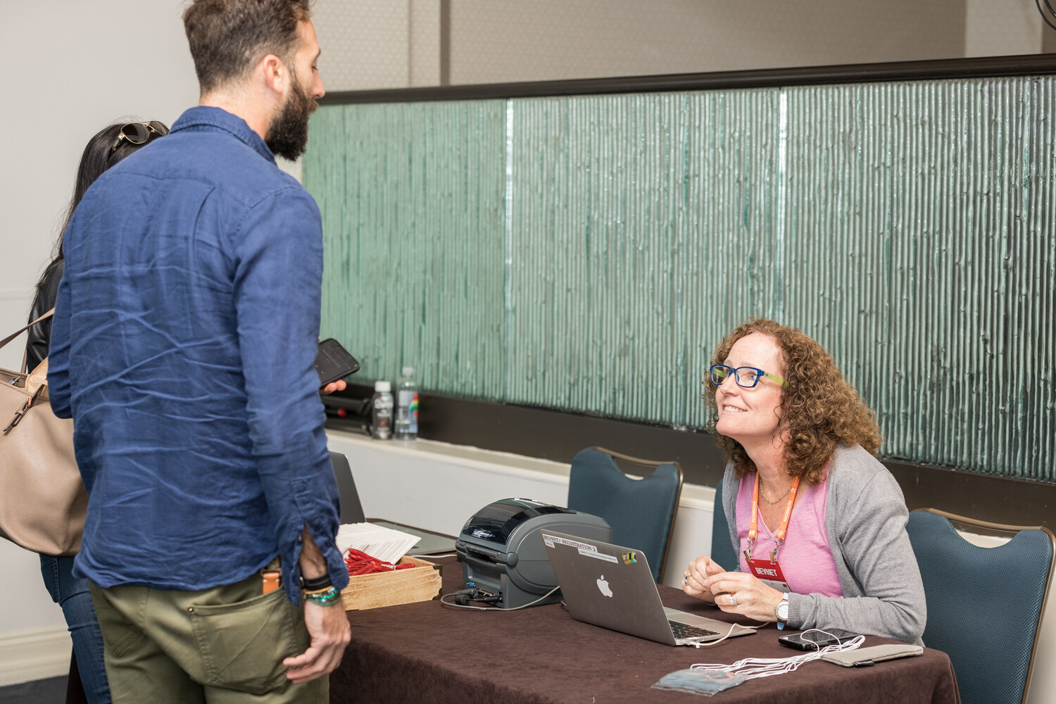 A bearded man in a blue linen shirt talks with a smiling seated woman with curly hair and glasses at a registration table with a laptop, badge printer and lanyards.
