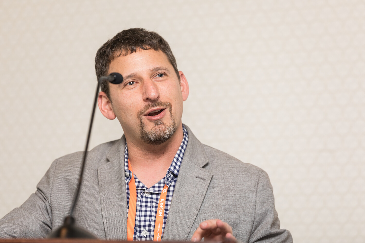 A man with a goatee in a gray blazer and navy gingham shirt with an orange lanyard speaks into a lectern microphone against a beige patterned wall.