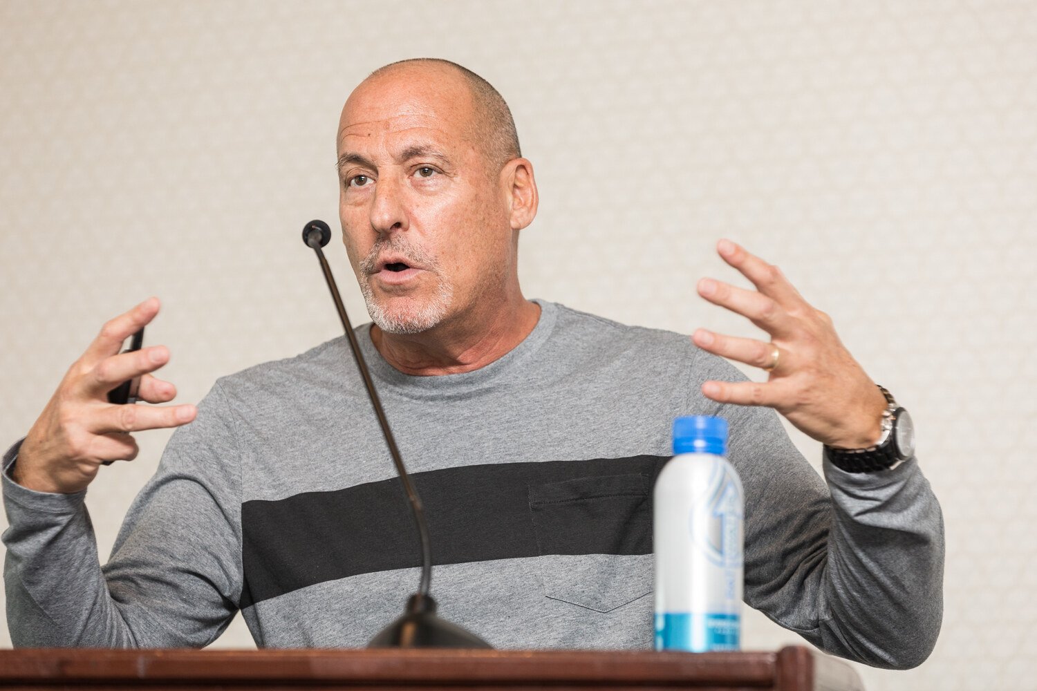 A bald man in a gray sweater with a black stripe holds both hands up while speaking at a wooden lectern, with a blue-capped water bottle beside the microphone.