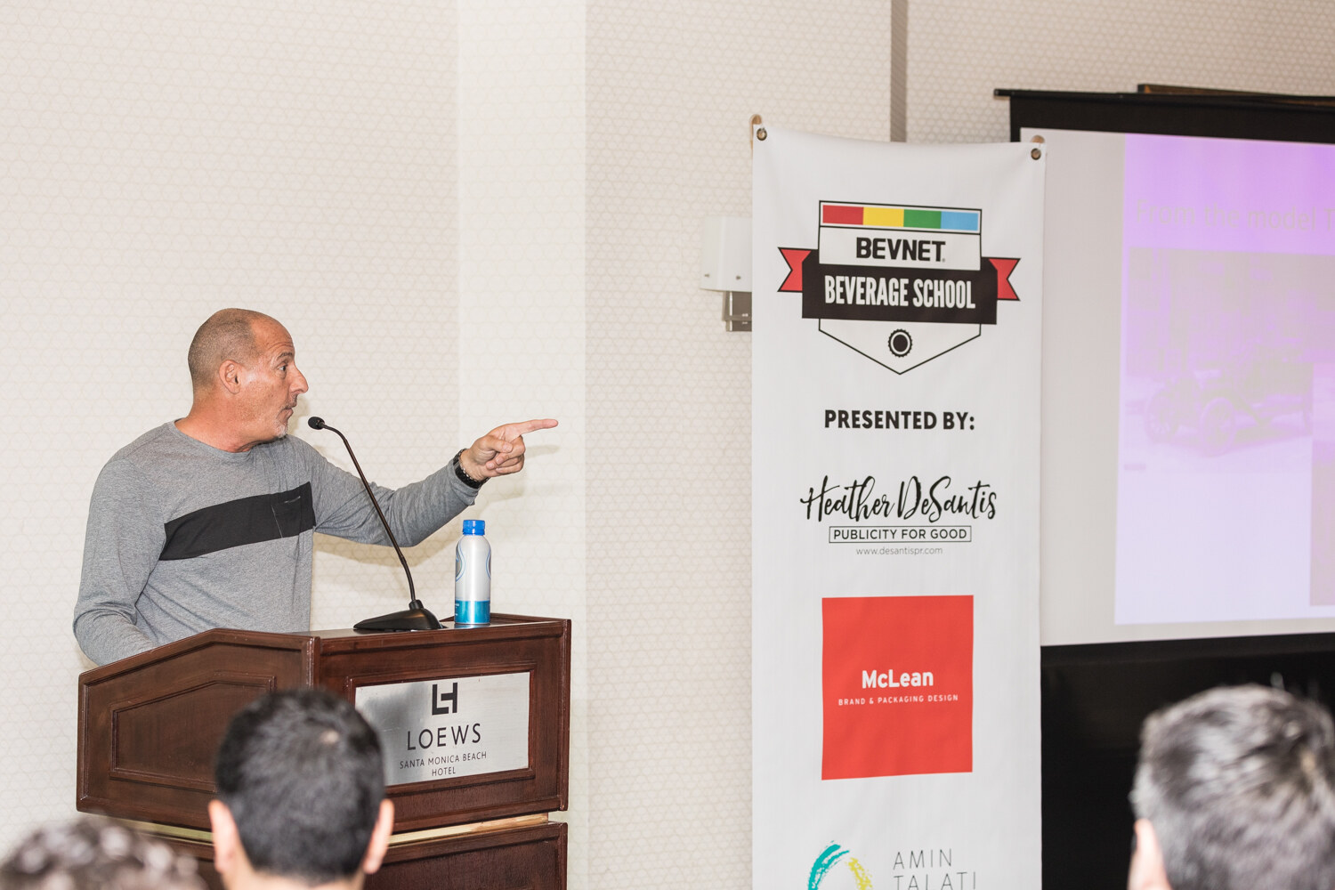 A man in a gray sweater stands at a wooden Loews lectern pointing toward a screen, beside a banner reading BevNET Beverage School, presented by Heather DeSantis Publicity for Good, McLean and Amin Talati.