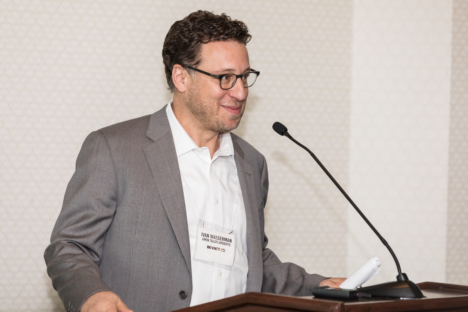 A man with curly dark hair and round glasses in a gray blazer and white shirt smiles beside a lectern microphone in front of a beige patterned wall.
