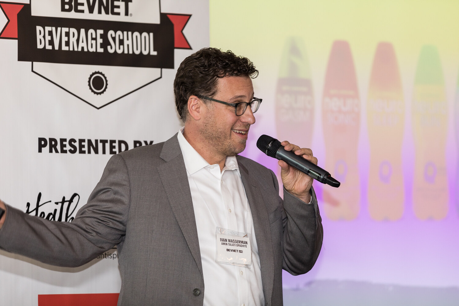 A man in a gray blazer and white shirt holds a handheld microphone and extends one arm in front of a BevNET Beverage School banner and a screen showing blurred colorful beverage bottles.
