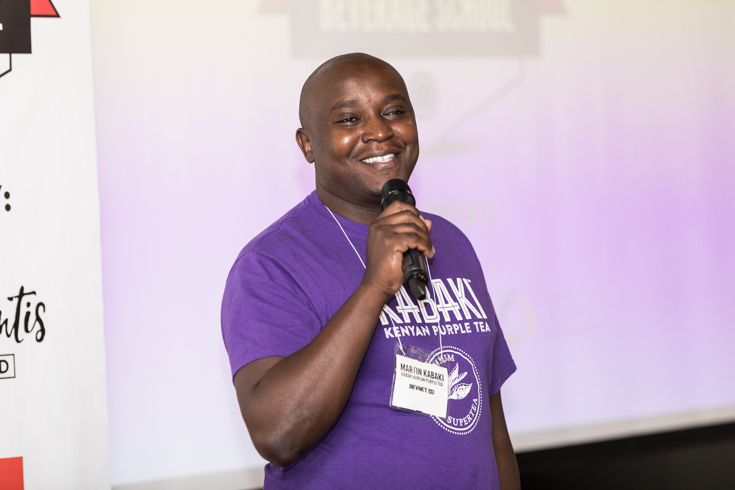 A smiling bald man in a purple Kabaki Kenyan Purple Tea T-shirt holds a handheld microphone in front of a projection screen.