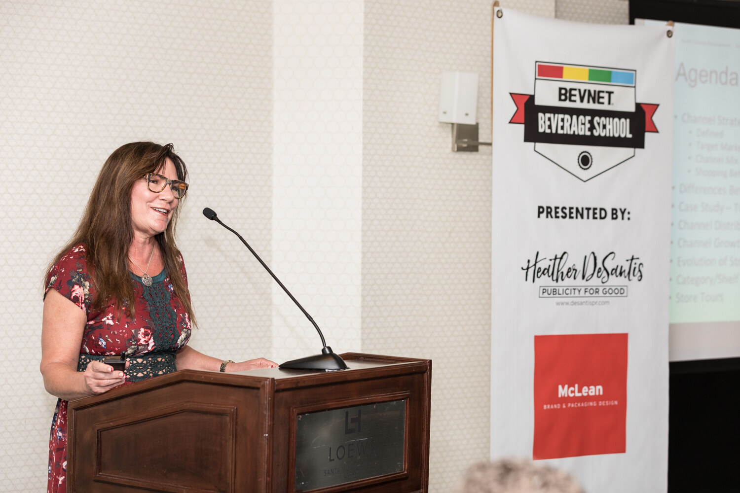 A woman with long brown hair and glasses in a red floral top speaks at a wooden lectern beside a banner reading BevNET Beverage School and a screen showing an agenda.
