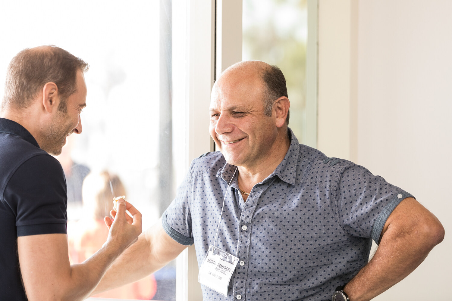 A man in a blue patterned shirt with a lanyard smiles and talks with a man holding a small snack in front of a bright window.