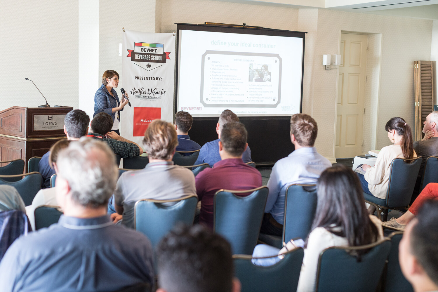 View over the heads of seated attendees toward a woman holding a microphone beside a Beverage School banner and a projector screen showing a slide titled define your ideal consumer.