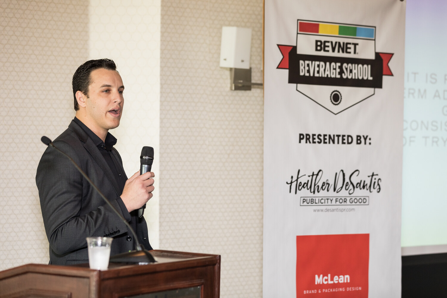 A man in a dark blazer and black shirt holds a handheld microphone beside a wooden lectern with a water cup, in front of a BevNET Beverage School banner.