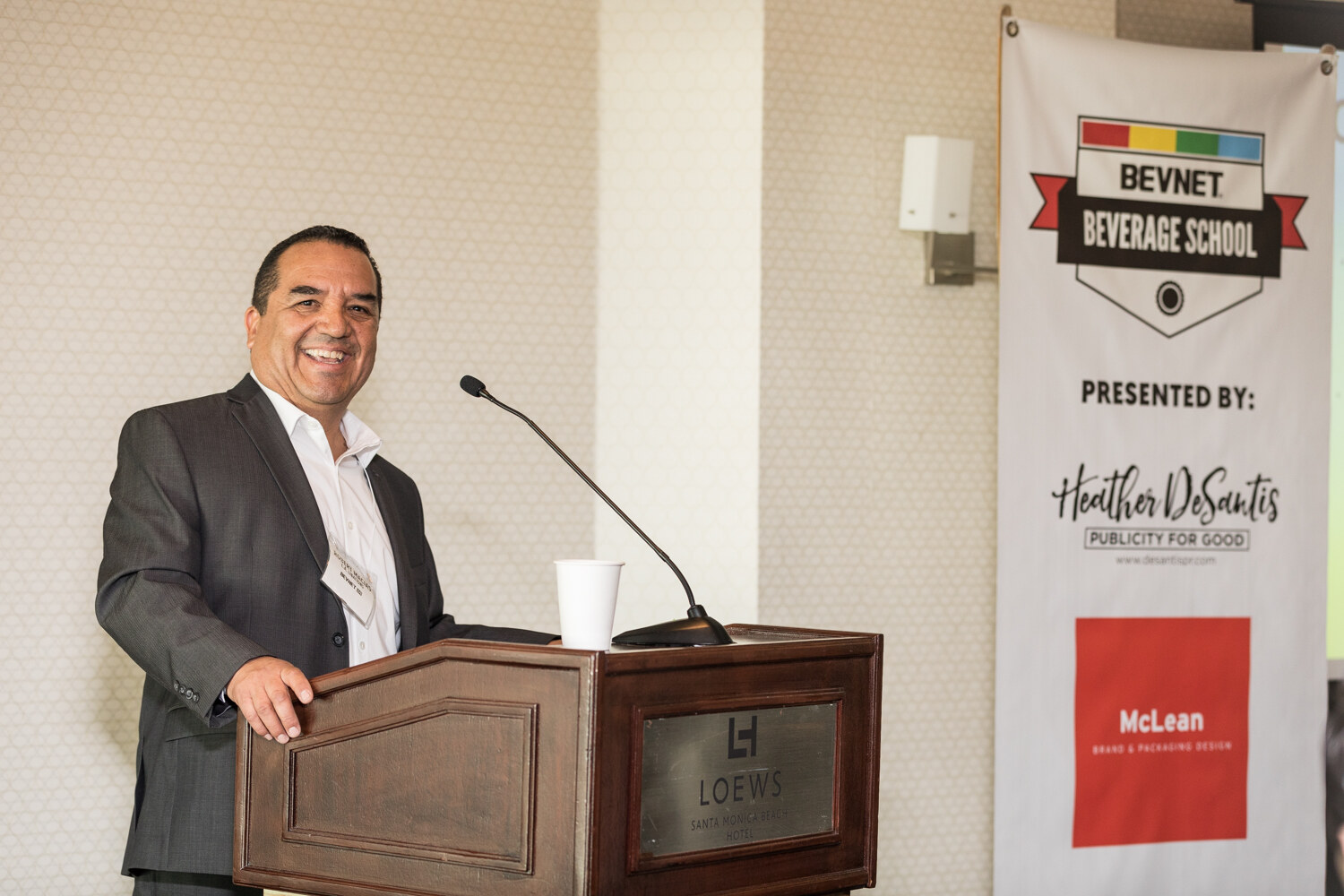 A smiling man in a gray blazer and white open-collar shirt leans on a wooden Loews Santa Monica Beach Hotel lectern next to a BevNET Beverage School banner.