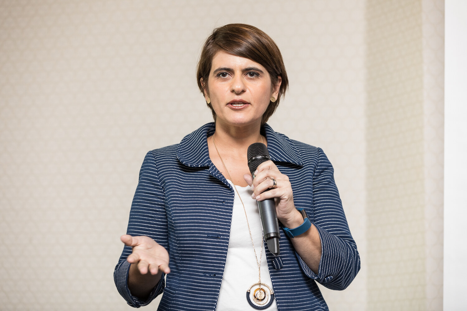 A woman with short brown hair in a navy striped blazer and white top holds a handheld microphone and extends her other hand while speaking.