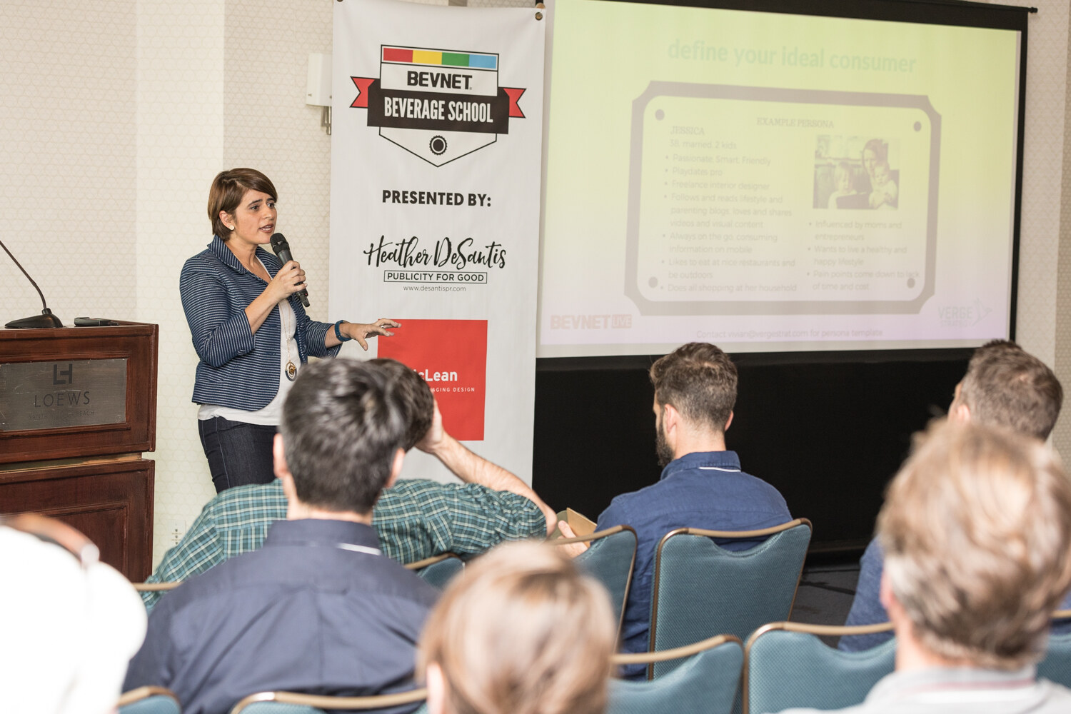 A woman in a navy striped blazer holds a microphone beside a BevNET Beverage School banner and a screen reading define your ideal consumer, with attendees seated in rows facing her.