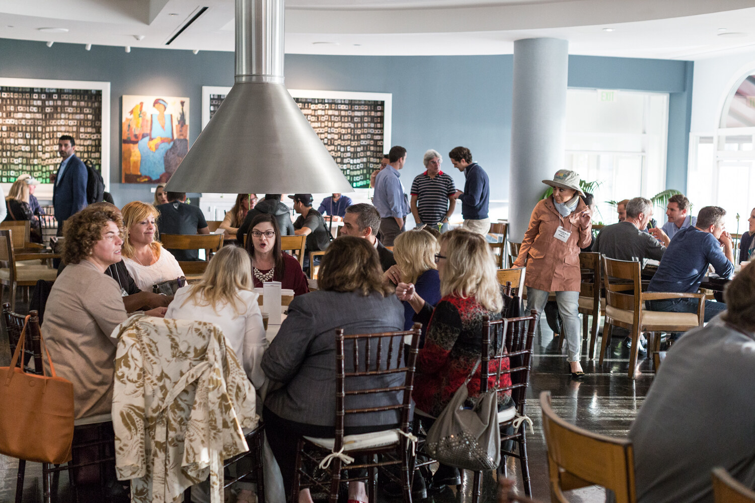 Wide view of a dining room where people sit at wooden tables and stand talking beneath a stainless steel hood, with colorful paintings on blue walls.