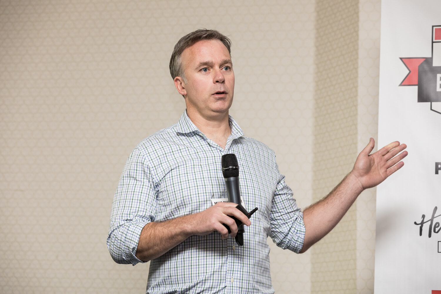 A man in a rolled-sleeve checked shirt holds a handheld microphone and clicker and opens one hand while speaking in front of a beige patterned wall.