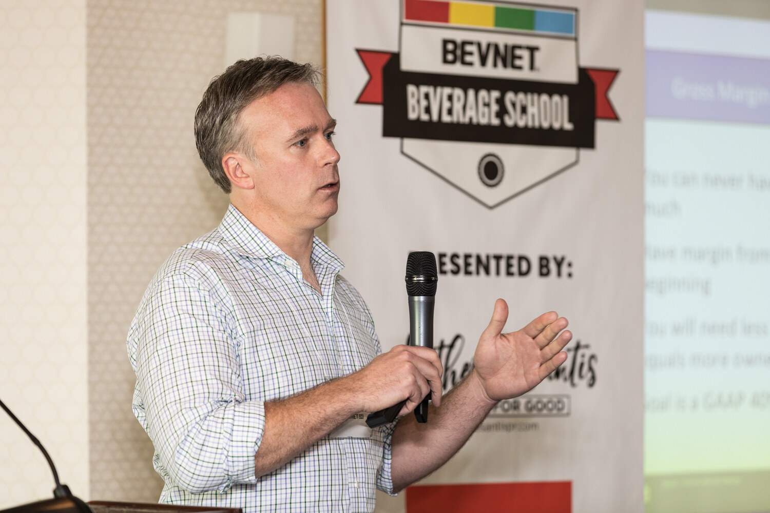 A man in a checked shirt holds a microphone beside a BevNET Beverage School banner with a slide on gross margin partly visible at right.