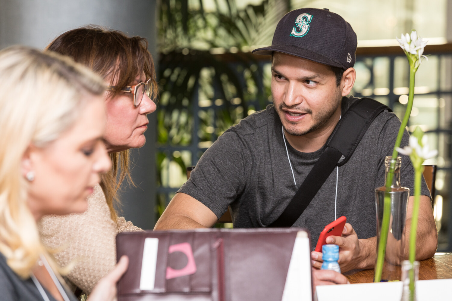 A man in a navy baseball cap and gray T-shirt holding a red phone talks with two women in the foreground at a wooden table with a white flower in a glass vase.