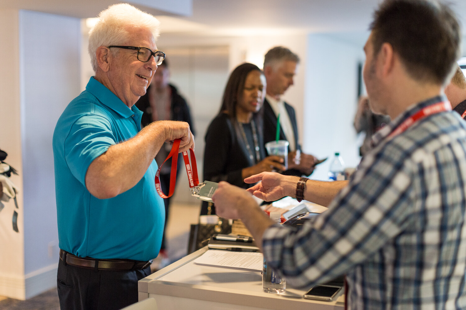 A smiling older man in a teal polo hands a red lanyard to a man in a plaid shirt at a registration counter with other attendees waiting behind.