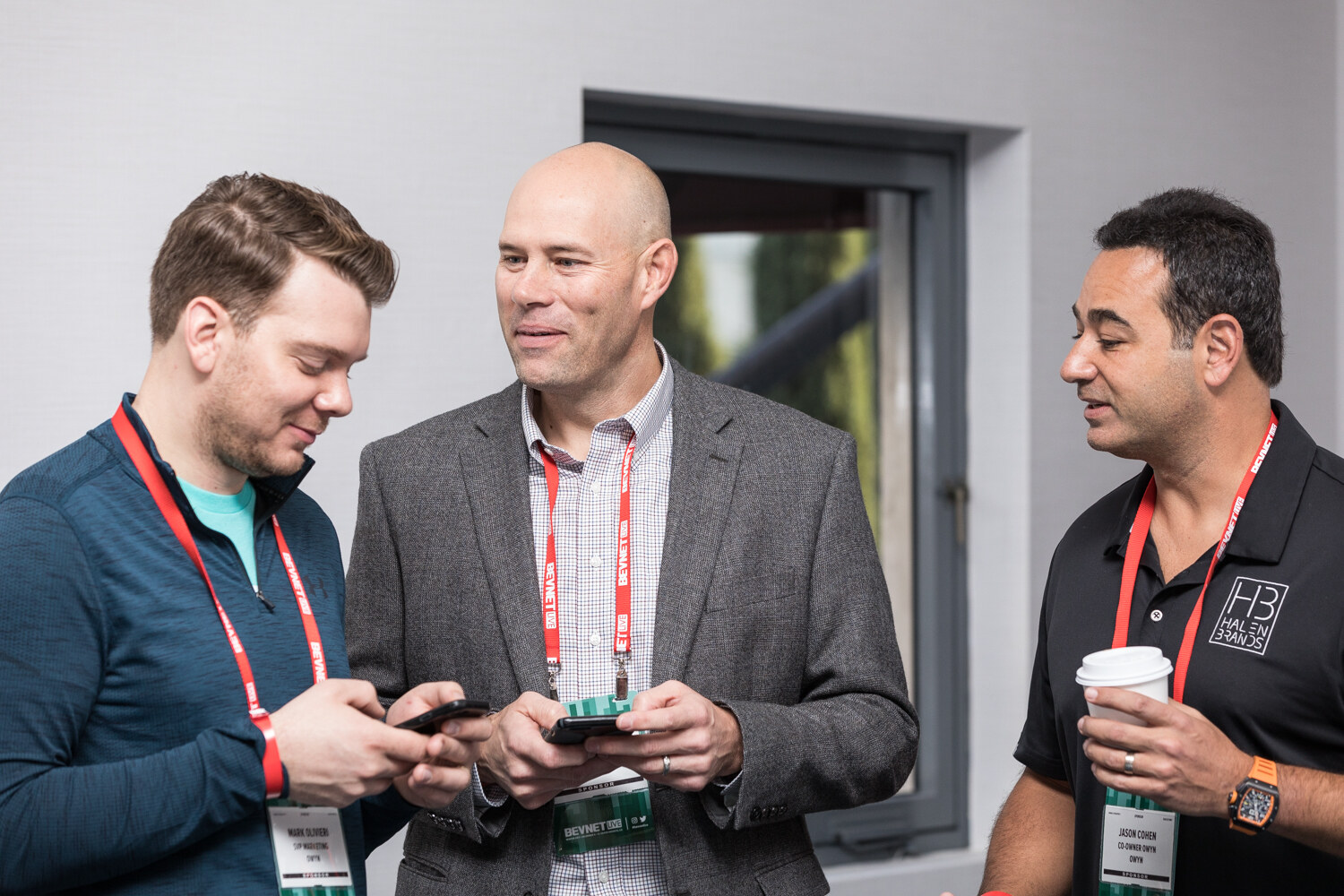 Three men with red lanyards stand in a hallway, two looking at phones and one in a black polo holding a coffee cup.