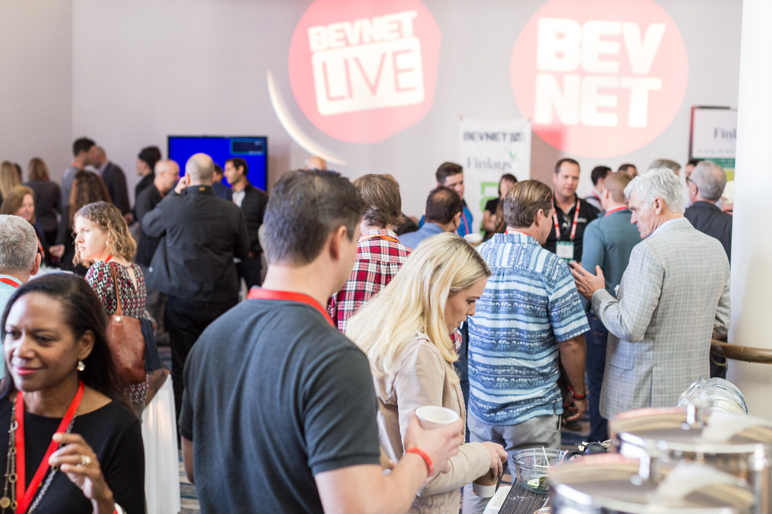 A crowded room of conference attendees with red lanyards talking, with large projected BevNET Live and BevNET circular logos on the wall behind them.