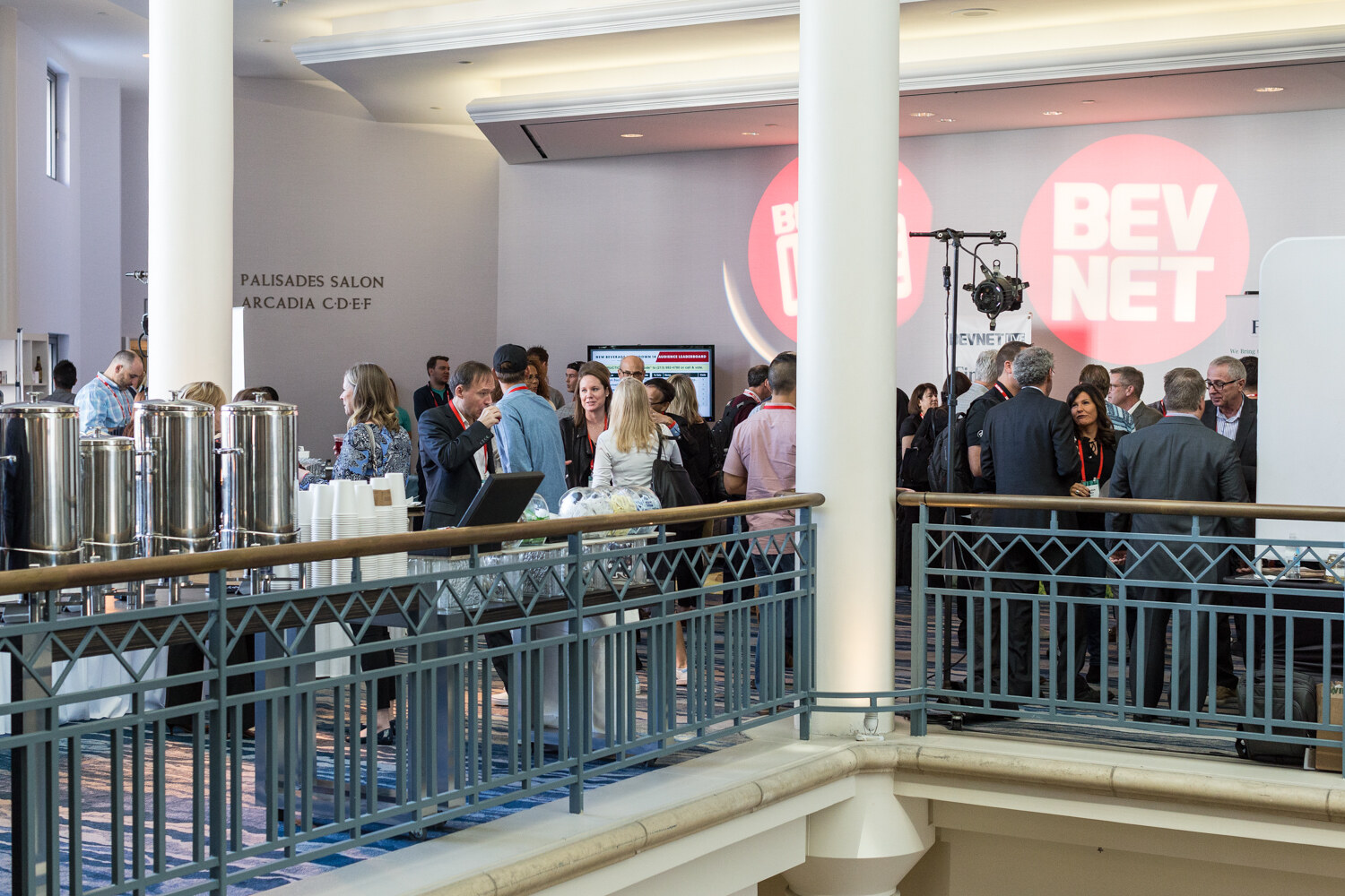 View from a balcony railing of attendees gathered near a row of stainless coffee urns and a wall with projected BevNET logos and a sign reading Palisades Salon, Arcadia.