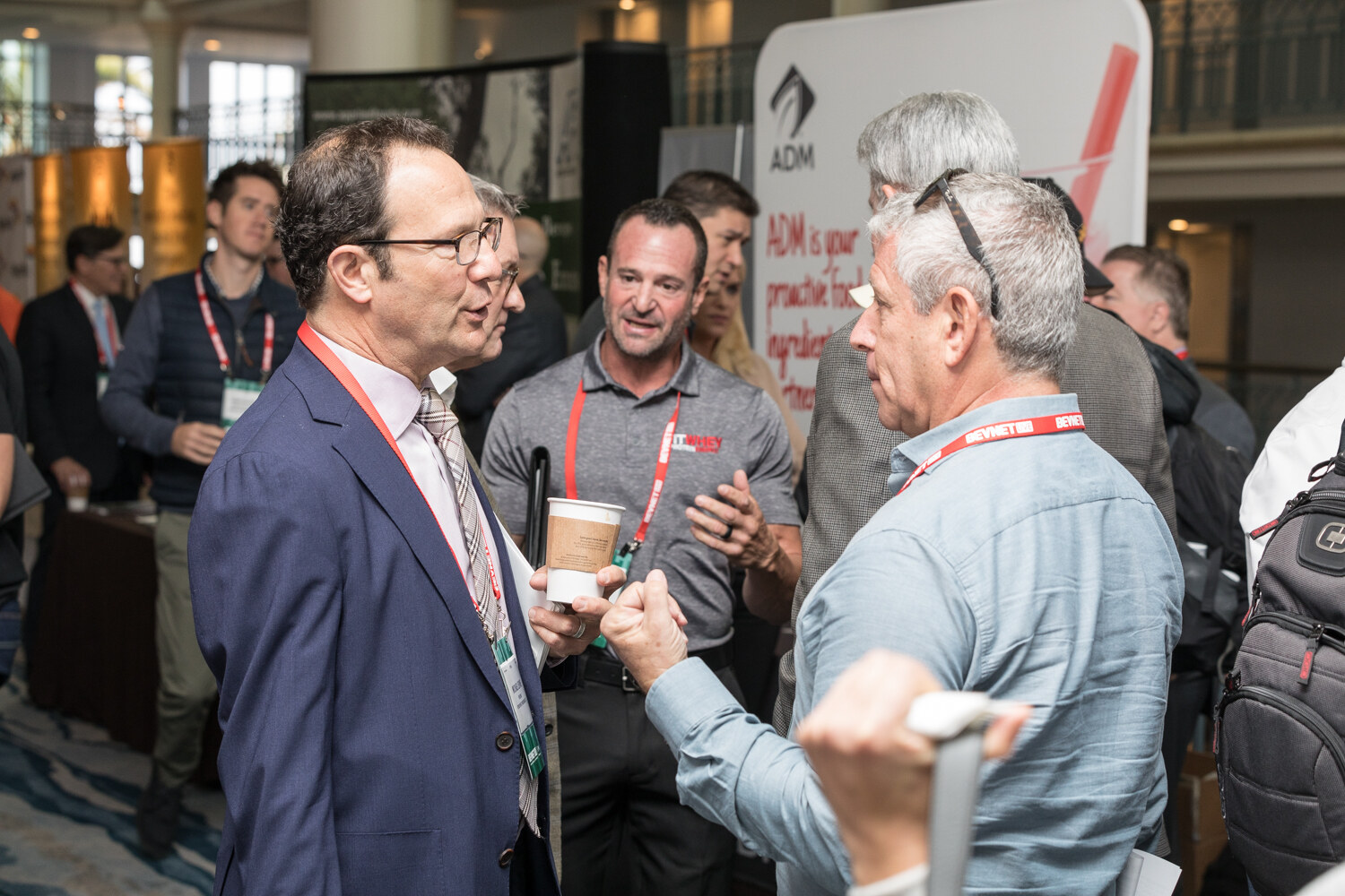 A small group of attendees with lanyards and coffee cups talks beside a banner reading ADM is your proactive food ingredient partner.