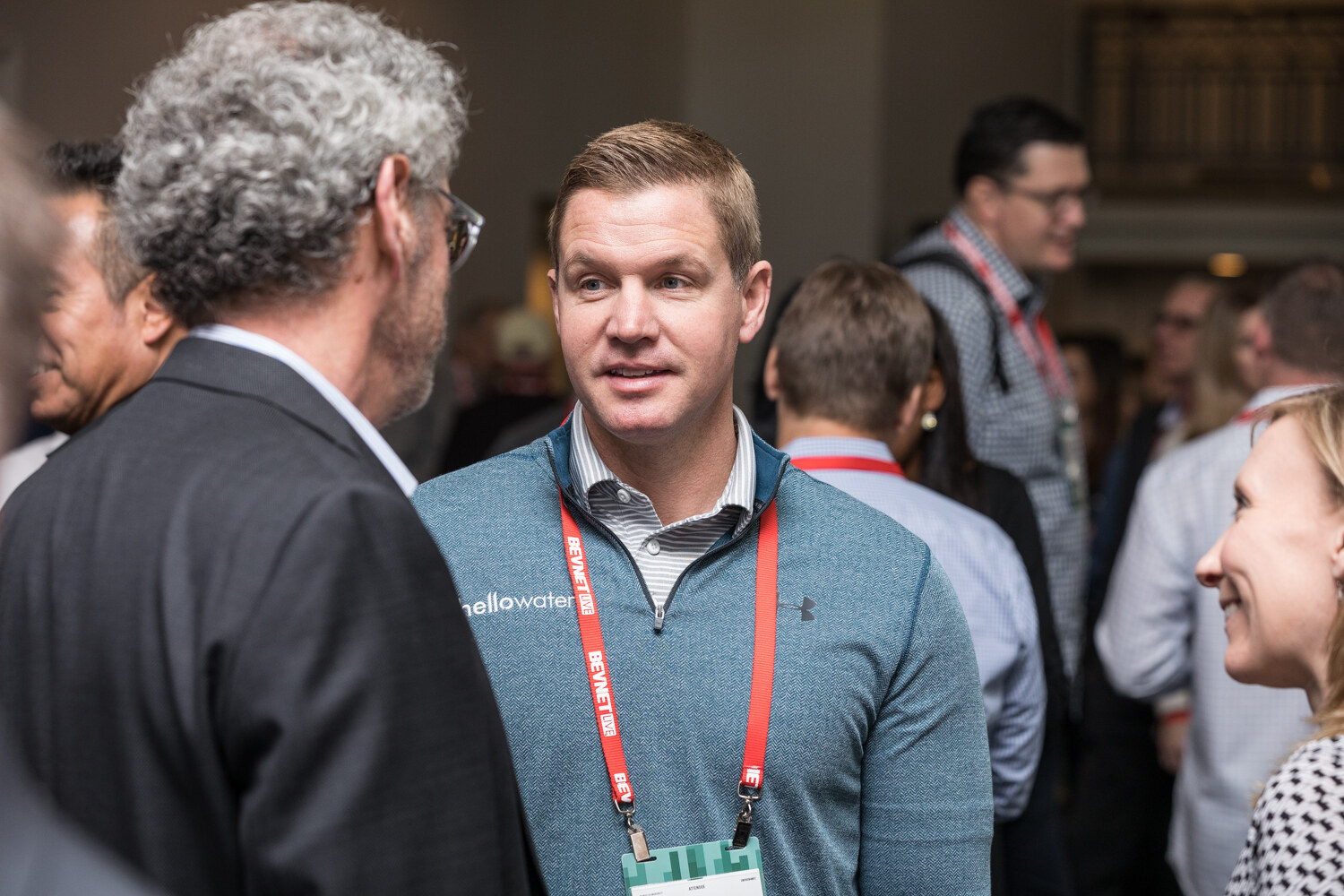 A man in a teal Under Armour pullover with a BevNET lanyard talks with a curly gray-haired man in a dark blazer, with other attendees in the background.