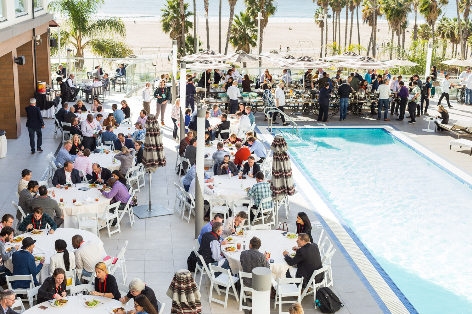 View from above of attendees at white round tables and a buffet line on a pool deck, with a blue pool at right and a sandy beach with palm trees in the distance.