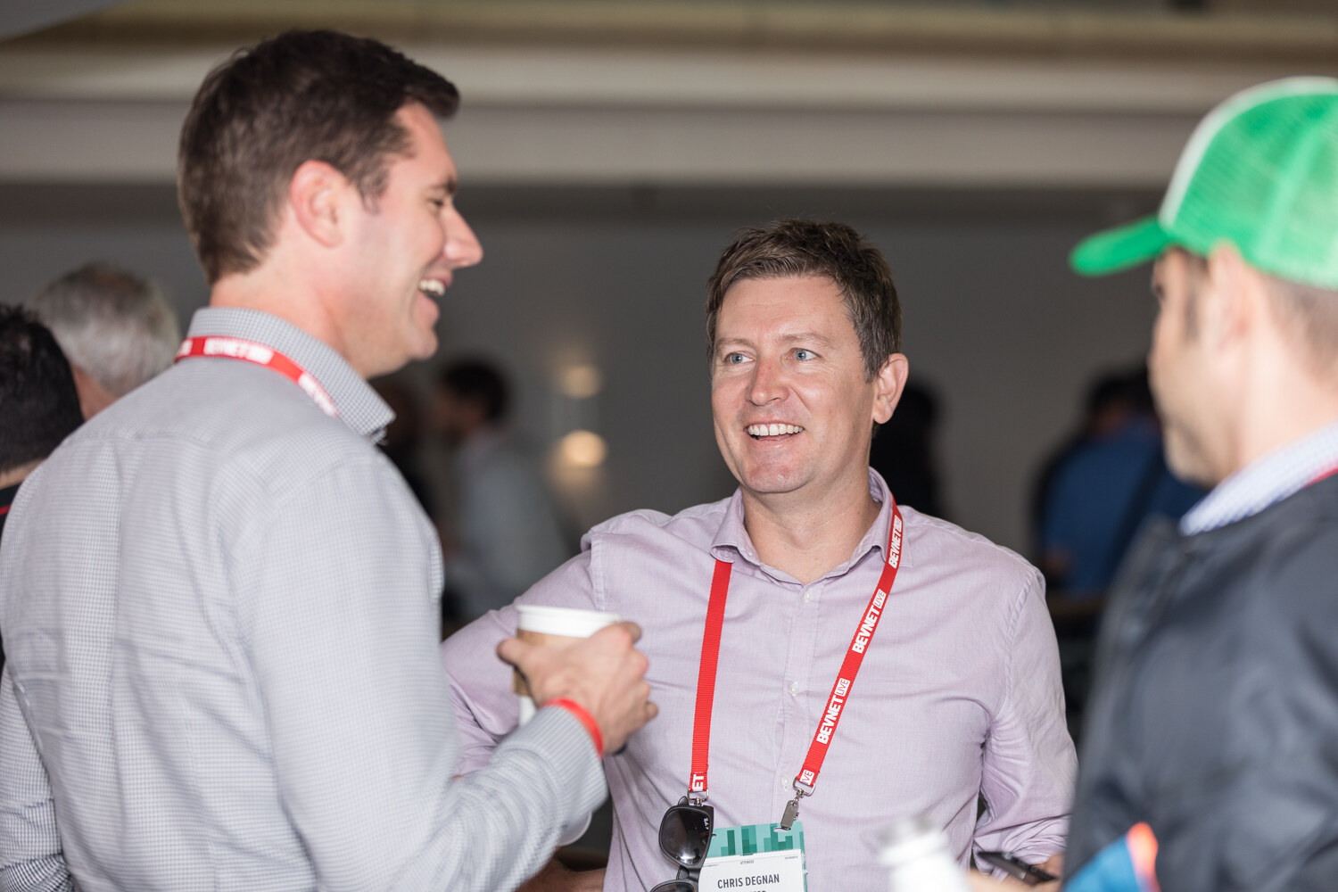 A smiling man in a lavender shirt with a BevNET lanyard talks with a man holding a coffee cup, with a person in a green cap at the edge of the frame.
