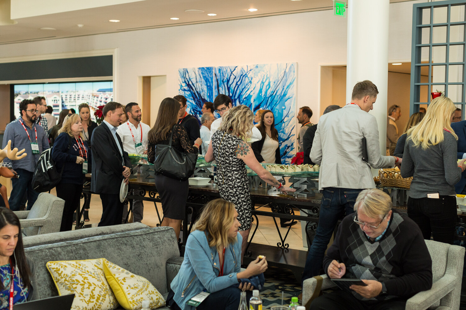 Attendees stand at a long buffet table in a hotel lobby with a large blue abstract painting behind them, while people sit on gray sofas in the foreground.