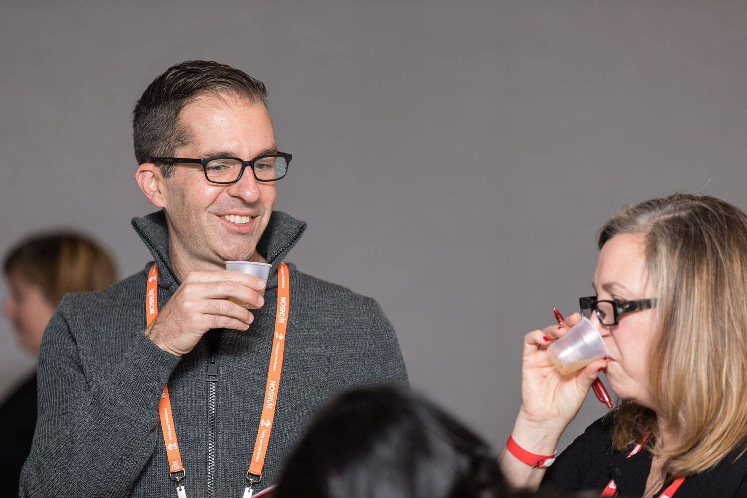 A smiling man with glasses and a gray zip sweater holds a small plastic cup while a woman with glasses beside him drinks from hers, both wearing orange lanyards.