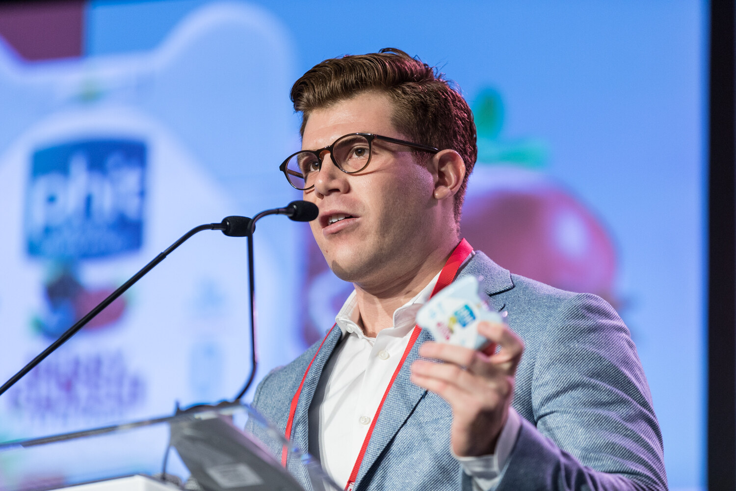 A man in round glasses and a light gray blazer speaks at a lectern with two microphones, holding a small colorful package, with a blurred slide showing the phit logo behind him.