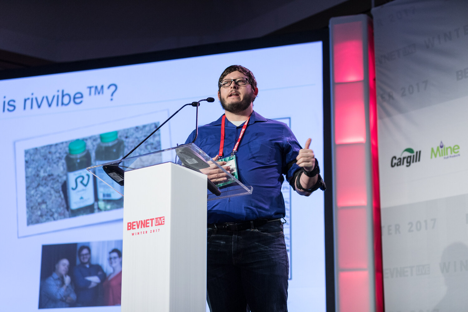 A bearded man in a blue shirt and glasses gives a thumbs up at a white lectern, beside a slide with bottles and the words Who is rivvibe.
