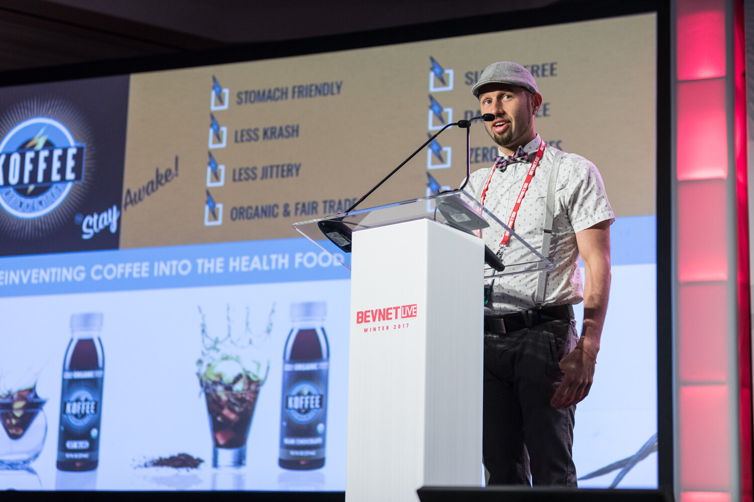 A man in a gray flat cap, bow tie and suspenders speaks at a white lectern beside a large slide for Koffee listing stomach friendly, less krash, less jittery and organic and fair trade.