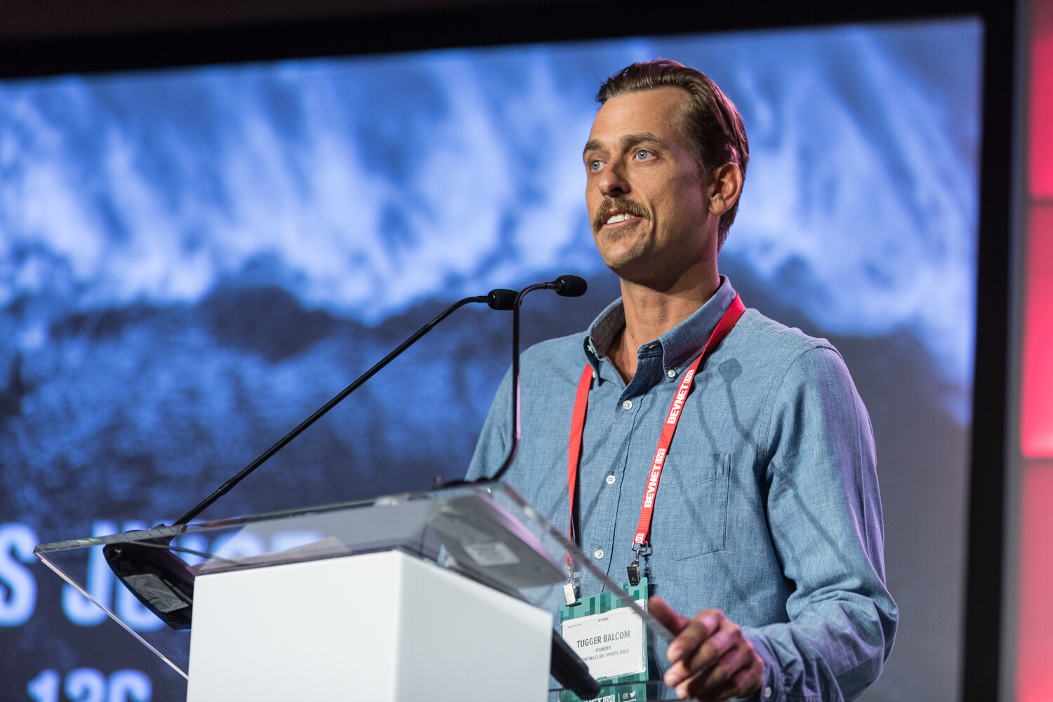 A man with a mustache in a blue chambray shirt and red lanyard speaks at a lectern with two microphones in front of a blue wave-patterned slide.