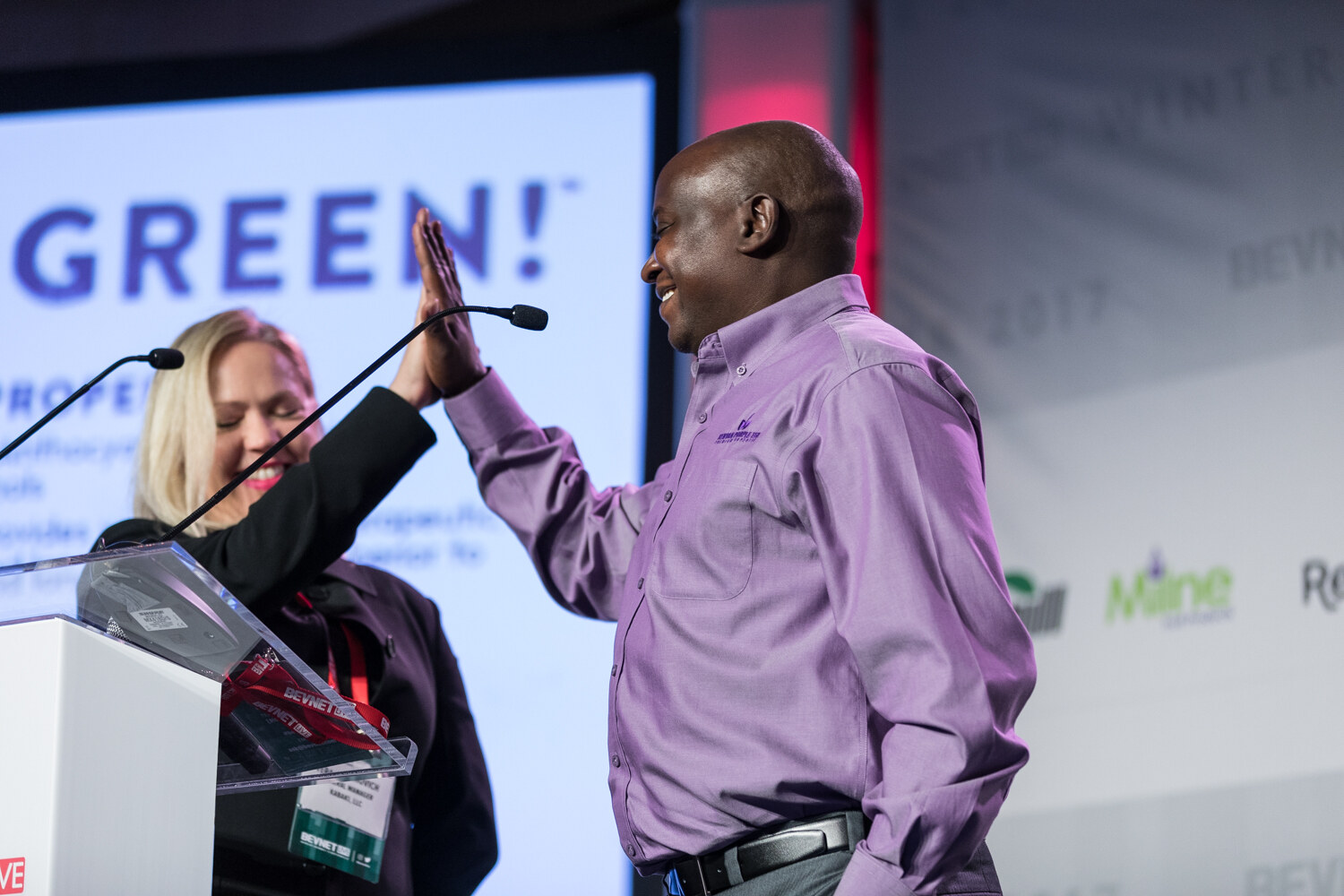 A smiling blonde woman and a man in a lavender button-up shirt slap hands in a high five at a lectern, with a bright slide reading Green behind them.