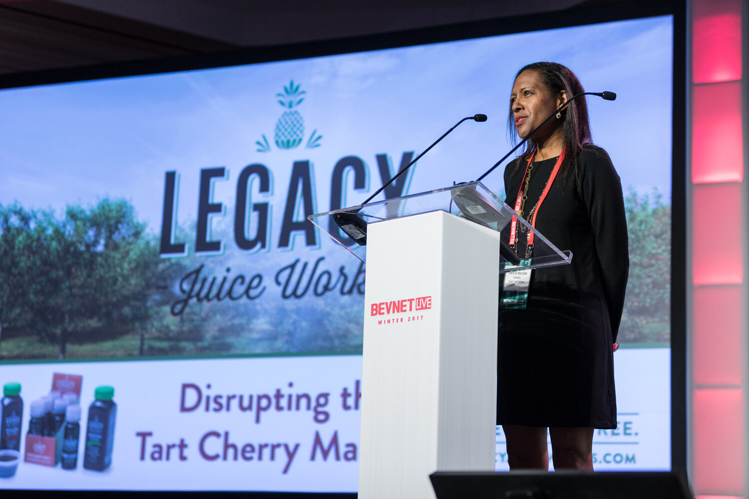A woman in a black dress stands at a white lectern with two microphones in front of a screen reading Legacy Juice Works, Disrupting the Tart Cherry Market.