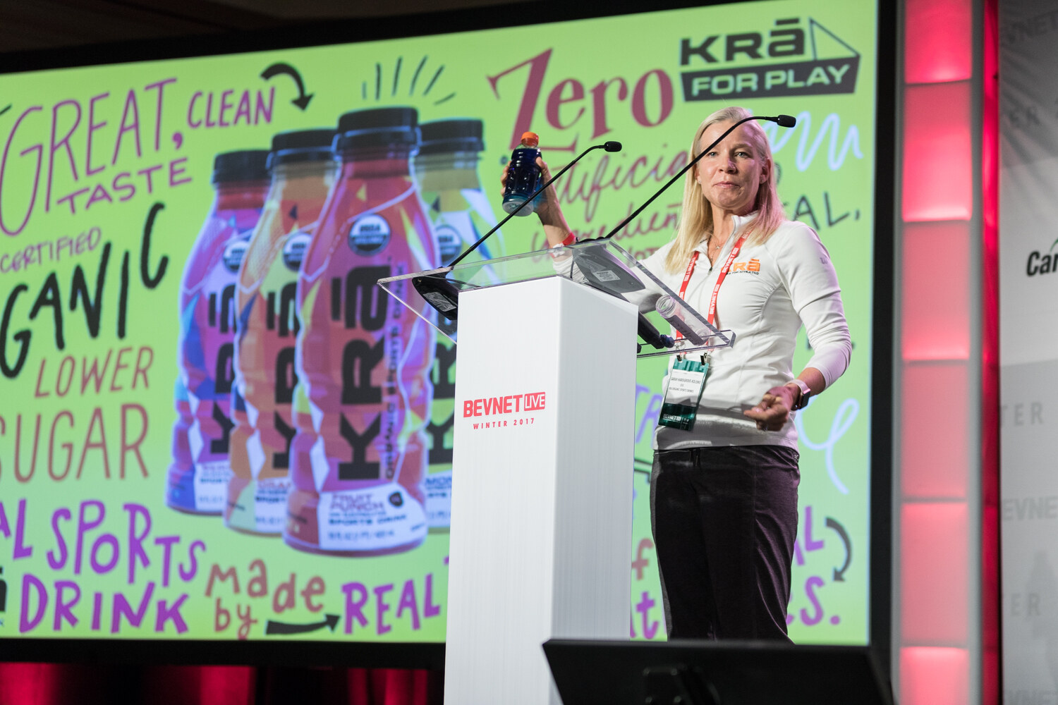 A blonde woman in a white top holds up a bottle at a white lectern in front of a green slide reading Great taste, certified organic, lower sugar, with KRA For Play bottles.