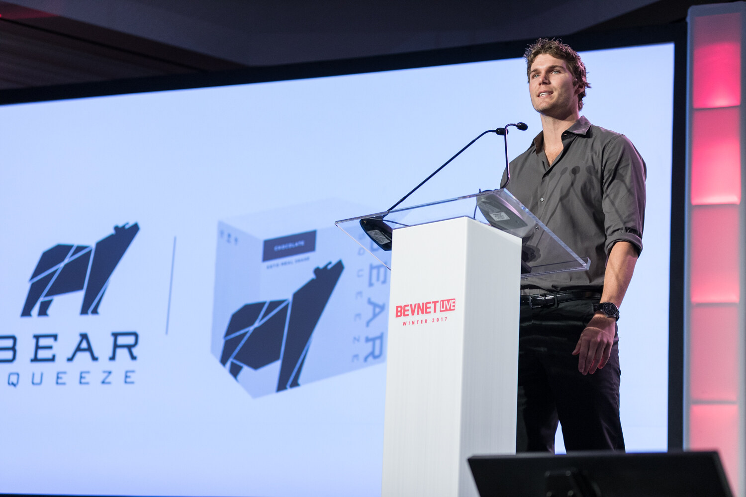 A man in a rolled-sleeve gray shirt stands at a white lectern in front of a slide showing the Bear Squeeze bear logo and a chocolate product box.