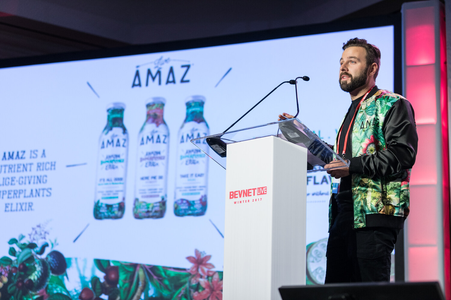 A bearded man in a green floral bomber jacket stands at a white lectern next to a slide showing three AMAZ bottles and the words AMAZ is a nutrient rich superplants elixir.