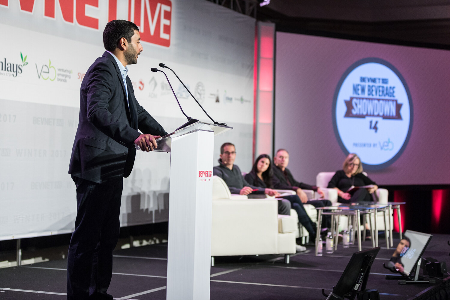 A man in a dark blazer speaks at a lectern with microphones while four judges sit on white sofas in the background, with a screen showing the BevNET New Beverage Showdown 14 logo.