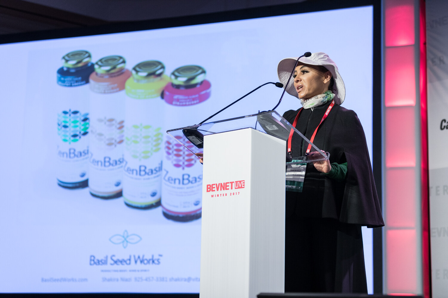 A woman in a white wide-brim hat and black cape speaks at a white lectern beside a slide showing four ZenBasil bottles and the Basil Seed Works logo.